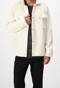 Vit fleecetröja med en texturerad yta, tryckknappsstängning, bröstfickor och en strukturerad krage, bärs över en mörk tröja och svarta byxor.