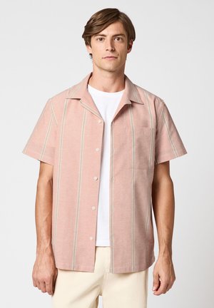 Homme portant une chemise rose à manches courtes avec des rayures verticales sur un t-shirt blanc et un pantalon crème, debout devant un fond uni.