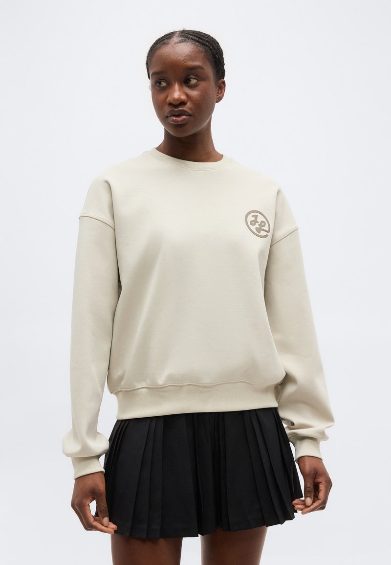 Femme portant un sweat-shirt beige clair avec un petit logo sur la poitrine et une jupe plissée noire, debout devant un fond uni.