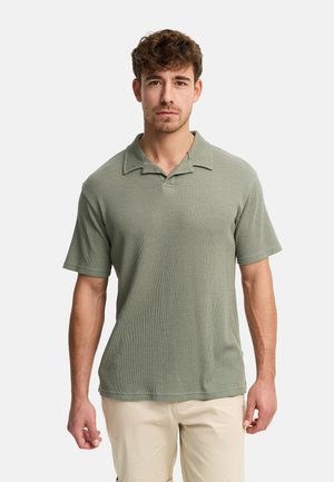 Mand iført en grøn kortærmet skjorte med krave og beige bukser, stående mod en ensartet lys baggrund.