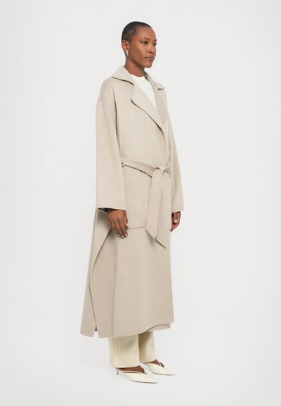 Cappotto avvolgente beige in lana con ampio risvolto e cintura, caratterizzato da maniche lunghe e una vestibilità morbida, indossato sopra pantaloni chiari e scarpe a punta.