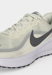 Λευκό και γκρι παπούτσι τρεξίματος Nike με πλέγμα στο άνω μέρος, λευκά κορδόνια, μαύρο λογότυπο Swoosh και παχιά λευκή σόλα, εμφανίζονται από πλευρική γωνία.