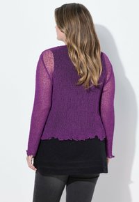 Lila stickad cardigan med en texturerad design, långa ärmar och en vågig nederkant. Buren över en svart topp och grå byxor.