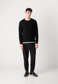 Svart ribbad tröja med långa ärmar som är lagd över en vit crewneck-tröja, matchad med svarta avsmalnande sweatpants och mörka sneakers.