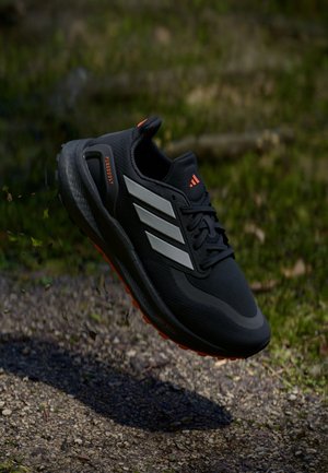Chaussure de course noire avec des rayures grises et des accents orange flottant au-dessus d'un sentier de gravier en plein air avec de l'herbe verte en arrière-plan.
