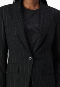 Černý pruhovaný blazer s přiléhavým střihem, zapínáním na jeden knoflík a strukturovanou tkaninou. Nositý přes černou košili s dekorativním vzorem.