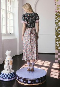 Žena stojaca na vyzdobenej kruhovej platforme, oblečená v kvetinových šatách s miešanými vzormi, levanduľovými ponožkami a béžovými lodičkami v slnečnej miestnosti.
