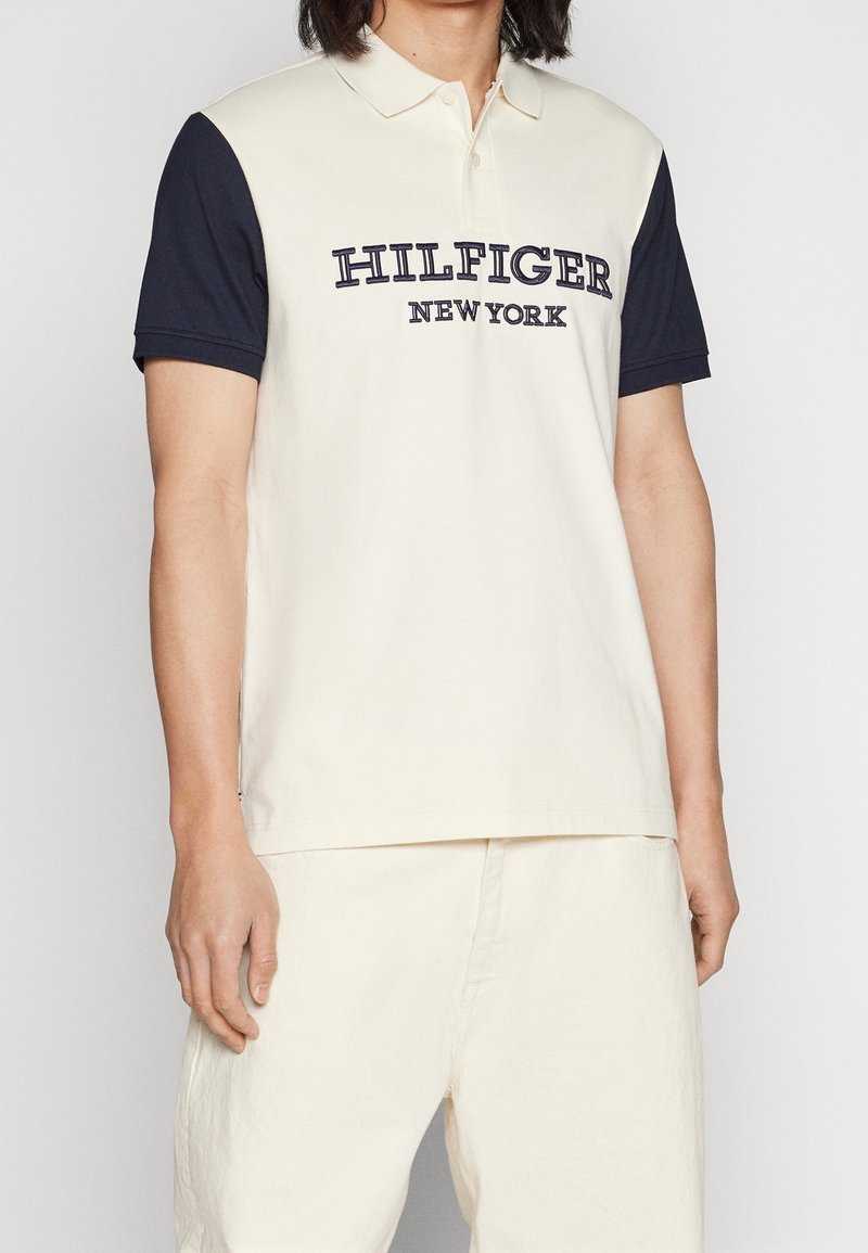 Personne portant un polo blanc avec des manches noires et le texte "HILFIGER NEW YORK" brodé sur le devant.