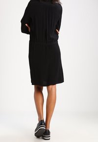 Czarna sukienka z wcięciem w talii, długimi rękawami i luźnym krojem. W zestawieniu z czarnymi butami z białymi paskami na podeszwie.