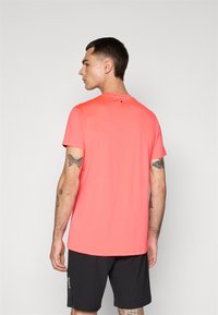 Mann som står med ryggen til iført en lys korallfarget kortermet skjorte og svarte shorts med synlige arm tatoveringer mot en ensfarget bakgrunn.