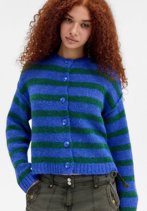 Jonge vrouw met krullend rood haar, gekleed in een blauwgroen gestreepte, dichtgeknoopte cardigan en donkergroene broek met handen in de zakken.