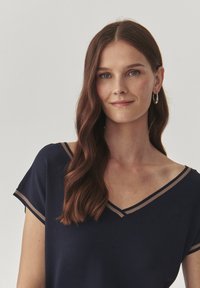 Laivastonsinisellä T-paitalla on lyhyet hihat ja V-pääntie, joka on varustettu kontrastivärisellä beigeillä reunuksilla kaula-aukon ja hihojen ympäri. Pehmeä kangas.