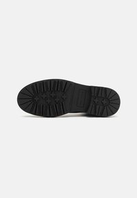 Black rubber shoe sole with deep tread pattern, featuring horizontal and vertical grooves. Label with brand name on the side.