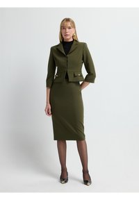 Olivgrön skräddarsydd kostym med en figursydd jacka med trekvartsärmar och en knälång blyertskjol. Guldknappdetaljer på jackan.