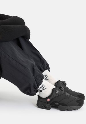 Person trägt schwarze Hosen und schwarze slip-on Reebok-Sneaker, kombiniert mit weißen Reebok-Crew-Socken vor einem einfarbigen weißen Hintergrund.