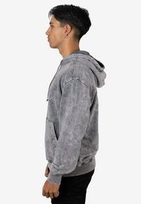 Sudadera gris con capucha, con un aspecto texturizado y desgastado, que incluye un bolsillo frontal y capucha con cordón. Puños y dobladillo acanalados. Vista lateral mostrada.