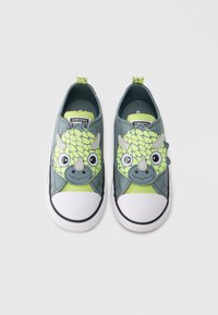 Πράσινα και γκρι πάνινα παπούτσια χωρίς κορδόνια με πρόσωπο δεινοσαύρου, αγκάθια, μεγάλα μάτια και λευκή λαστιχένια μύτη. Ζωντανές πινελιές τονίζουν το σχέδιο.