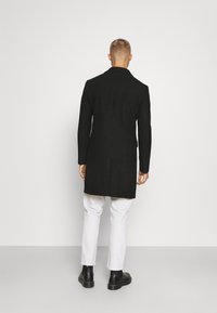 Cappotto di lana nera con una vestibilità su misura, dotato di un colletto a punte, maniche lunghe e taglio dritto. Indossato sopra pantaloni bianchi e stivali neri.
