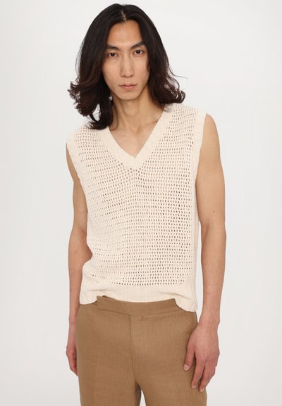 Débardeur en tricot beige clair sans manches avec un col en V et une texture ajourée, associé à un pantalon marron ajusté. Design simple et décontracté.