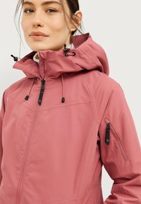 Giacca impermeabile rosa con colletto alto, cappuccio regolabile e tasca con zip sulla manica. Texture liscia e dettagli di design funzionali.