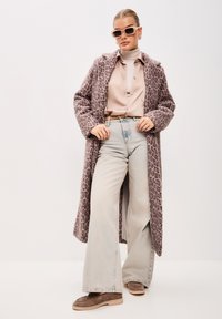 Lange jas met luipaardprint in paars en beige, beige blouse met knopen, lichtblauwe wijde jeans, grijze suède schoenen en roze zonnebril.
