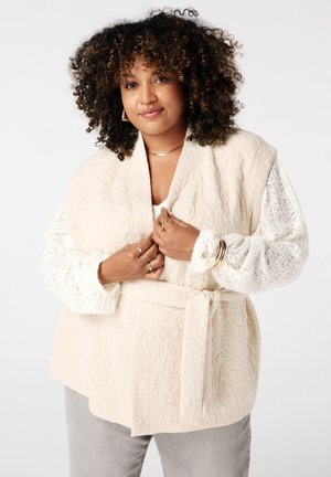 Crème gekleurde vest met een brede kraag en een strikceintuur, met mouwen afgewerkt met kant. Draagt over een witte top en grijze broek.