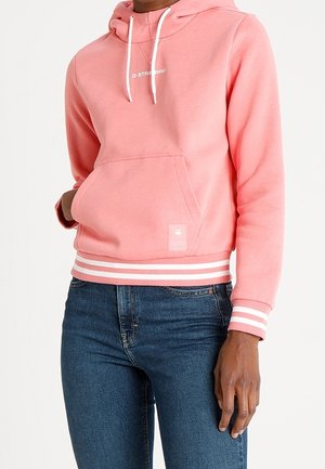 Personne portant un sweat à capuche rose avec des cordons blancs et des poignets rayés, associée à un jean bleu foncé, main reposant tranquillement sur le côté.