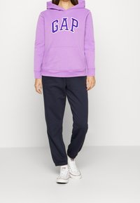Felpa viola con logo "GAP" in bianco e pantaloni jogger blu scuro. La felpa presenta una tasca a canguro e cappuccio regolabile con cordino.