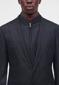 Gros plan d'une personne portant un blazer texturé foncé par-dessus une couche intérieure à col montant zippé, debout devant un fond clair uni.