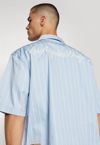 Camisa de manga curta azul clara com riscas verticais brancas e bordado "Los Angeles Dodgers" nas costas. Apresenta um colarinho reto e etiqueta.