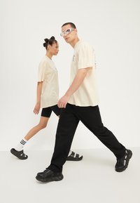 Due persone camminano fianco a fianco. Indossano t-shirt beige larghe e pantaloni neri. La donna ha pantaloni corti neri da ciclismo e sandali; l'uomo indossa sneaker nere.
