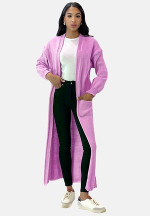 Lange roze cardigan met geribbelde textuur, open voorkant en zakken, gecombineerd met zwarte skinny jeans en witte sneakers.