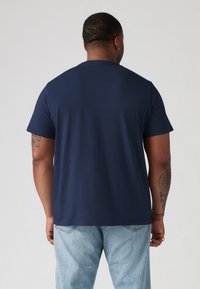 T-shirt en coton bleu marine à manches courtes, col rond et coupe droite. Présente une texture lisse et un design minimal, porté avec un jean bleu clair.