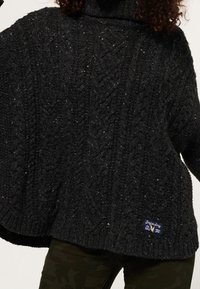 Zwarte chunky gebreide coltrui met kabelpatronen, gedragen over donkere camouflagebroek.