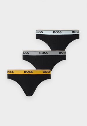 Young man standing barefoot, wearing black briefs with a yellow waistband labeled "BOSS," looking to the side against a plain white background.