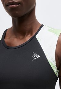 Κοντινό πλάνο ενός ατόμου που φοράει μαύρο αθλητικό τοπ με λευκές και ανοιχτοπράσινες σχεδιασμένες πλαϊνές λωρίδες στους ώμους και ένα μικρό λευκό λογότυπο στο στήθος.