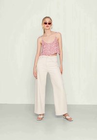 Top cropped floral cor-de-rosa com alças finas, combinado com calças largas de cor creme e sandálias brancas. Modelo a usar óculos de sol redondos.