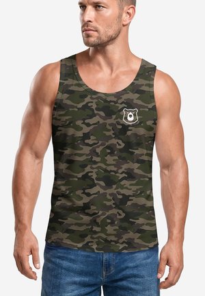 Muskelbepackter Mann, der ein grünes und braunes Tarn-Top mit kleinem weißem Bärenlogo auf der Brust trägt, kombiniert mit blauen Jeans.