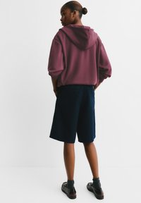 Weinrotes Kapuzensweatshirt mit einer Rückentasche, kombiniert mit marineblauen Shorts. Schwarze Schuhe mit sichtbaren grauen Socken am Knöchel.