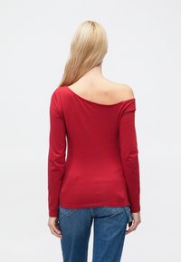 Červený top s dlouhým rukávem a odhalenými rameny. Vyrobený z hladké látky, má přiléhavý střih a minimalistický styl. Kombinován s modrými džíny.