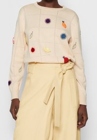 Kremowy sweter dziergany z haftowanymi owocami i kwiatowymi akcentami w różnych kolorach. Połączony z beżowymi spodniami o wysokim stanie.