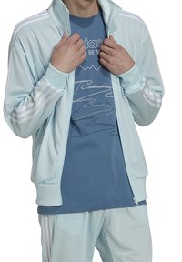 Lichtblauwe trainingsjack met witte strepen op de schouders, gecombineerd met een blauw T-shirt met een minimalistisch berggrafisch ontwerp.