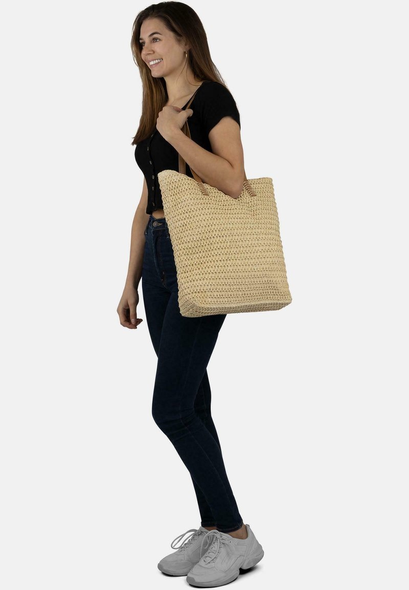 Geflochtene Strohtragetasche mit zwei Griffen, rechteckiger Form, beige Farbe und strukturierter Oberfläche. Frau hält die Tasche und trägt ein schwarzes Oberteil und Jeans.