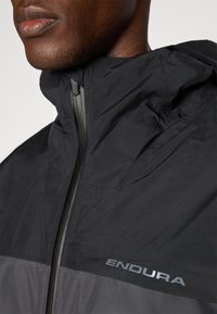 Veste coupe-vent noire avec des accents gris. Dispose d'un col haut, d'une fermeture éclair à l'avant et met en valeur le logo "ENDURA" dans une finition réfléchissante.