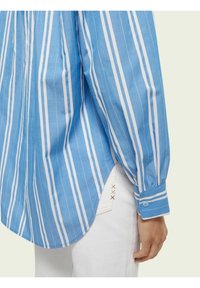 Blue and white striped long-sleeve shirt with a rounded hem and buttoned cuffs, made from lightweight fabric. Visible stitching detail on white pants.