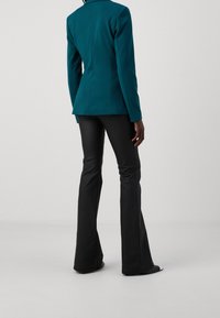 Person som bär en teal färgad kavaj och svarta utsvängda byxor, stående mot en enfärgad ljusgrå bakgrund, sedd bakifrån.