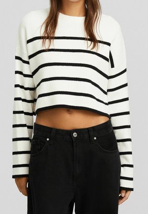 Kvinde iført en hvid cropped sweater med sorte vandrette striber og højtaljede sorte jeans, stående mod en ensfarvet baggrund.