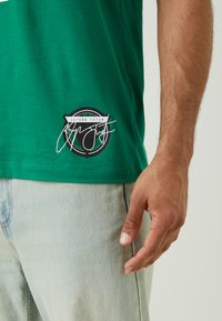 Green cotton T-shirt with a black and white circular logo featuring text and a signature. Light blue denim jeans partially visible.