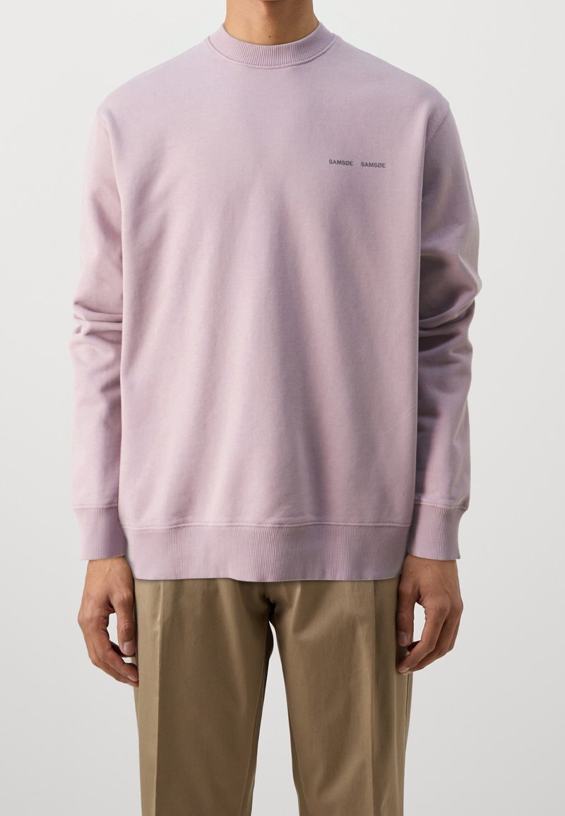 Personne portant un sweat-shirt à manches longues violet clair avec un petit texte "SAMSØE SAMSØE" sur la poitrine et un pantalon beige, debout immobile.
