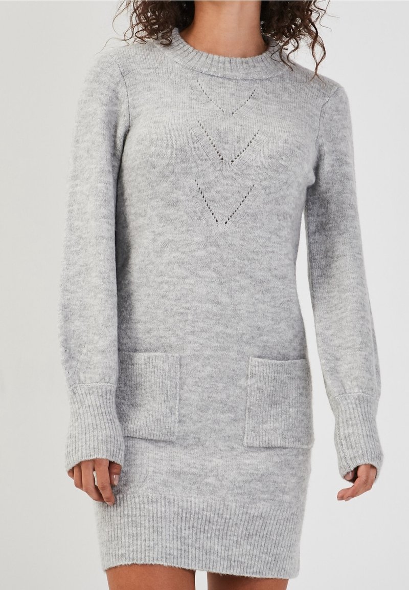 Femme portant une robe-pull en tricot gris clair à manches longues avec deux poches devant et un motif chevron sur la poitrine.